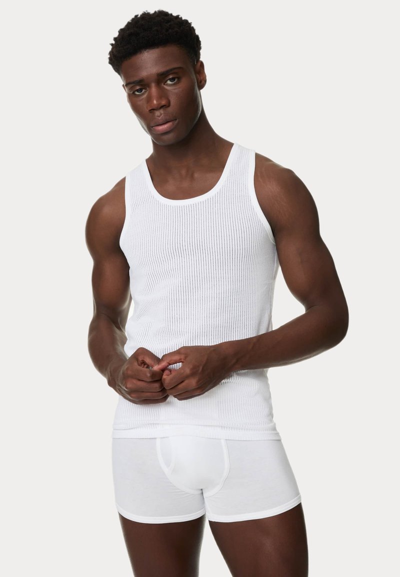 Weißes geripptes Tanktop und enge weiße Boxer-Briefs an einer Person mit neutralem Ausdruck, die glattes Material und körperbetonten Sitz zeigt.