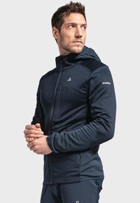 Navy blue softshell jacket with a hood, featuring zippered pockets, elastic cuffs, and contrasting shoulder panels. Minimal branding detail.
