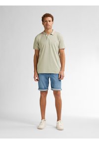 Lichtgroene polo met rode randen, gecombineerd met opgerolde denimshorts. Model draagt lichte sneakers en staat voor een eenvoudige achtergrond.