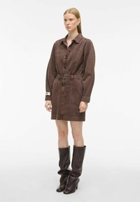 Smeđa traper haljina s naglašenim strukom, dugim rukavima, dugmadi sprijeda i dva prednja džepa. Uparena s smeškim čizmama do koljena.