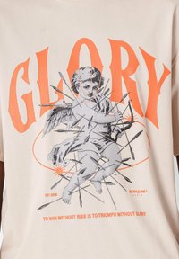 Beige t-shirt med kort ärm som har en grafik av en ängel med pilar och "GLORY" i fet orange text, med en ribbad textur.