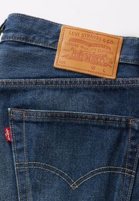 Blå denimjeans med en brun läderlapp som har varumärkeslogotyp och detaljer. Distinktiv gul sömnad och röd etikett på bakfickan.