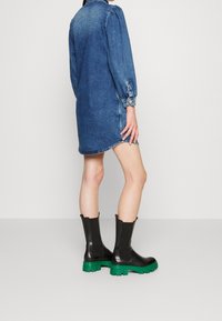 Personne portant une robe en jean bleu à manches longues et des bottines noires avec des semelles épaisses vert vif, debout devant un fond uni.