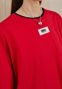 Person som bär en starkt röd sweatshirt med en vit MM6 Maison Margiela-logotyp och ett silverhalsband över en svart kragskjorta.
