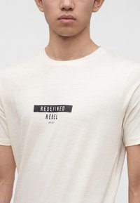Młoda osoba nosząca jasnobeżową koszulkę z czarnym napisem „REDEFINED REBEL EST 2013” na piersi.