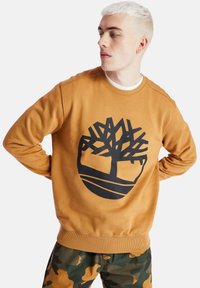 Felpa marrone con un grande logo di un albero nero sulla parte anteriore. Presenta un collo rotondo e polsini e orlo a costine. Abbinata a pantaloni mimetici.