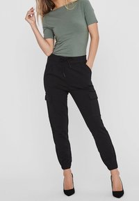 Olivově zelený krátký rukáv top spárovaný s černými cargo kalhotami s bočními kapsami a elastickými lemy, nošený s černými vysokými podpatky.