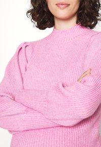 Light pink ribbed sweater with long sleeves, high neck, and puffed shoulder detail. Soft texture, fitted style with subtle pattern.
