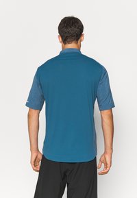 Homme portant une chemise à col à manches courtes bleu bicolore et un pantalon noir, se tenant de dos face à l'appareil photo.