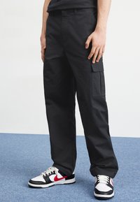 Svarta cargo-byxor i slitstark tyg, med en sidoficka, rak bendesign, och bärs med svartvita sneakers med röda detaljer.