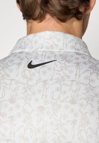Blomstermønstret Nike polo t-shirt med en hvid base og lysebeige designs. Sort Nike-logo på kraven. Blødt, letvægtsstof.