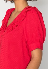 Blusa rossa con scollo arricciato, maniche a sbuffo corte e delicati pois testurizzati. Tessuto leggero con un design fluido.
