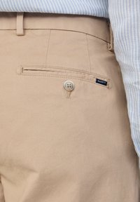 Pantaloni beige con una singola tasca posteriore con bottone e etichetta del marchio Hackett, indossati con una camicia a righe blu e bianche.