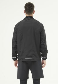Svart sportjacka med hög krage, långa ärmar och vita reflekterande detaljer. Slät, lätt material. Buren över svarta shorts.