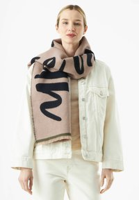 Roze wollen sjaal met zwarte letters, voorzien van een franjesrand en een groene accent aan de onderkant. Draagt over een crème kleurige top en jas.