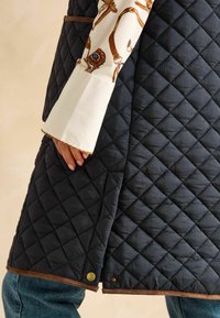 Jumta melna jaka ar brūnu akcentu. Satur dimanta formas šuvju, aizdari ar pogām un gludu faktūru. Redzama balta rakstota piedurkne.