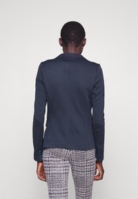 Person wearing a fitted dark navy blazer and black-and-white patterned pants, standing with their back to the camera on a plain background.