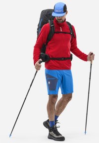 Veste en polaire rouge, shorts bleus avec une poche marine, bottes de randonnée noires, sac à dos gris et bâtons de trekking. Casquette bleue et noire.