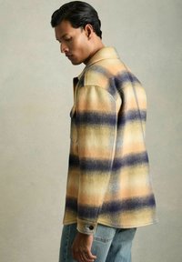 Camisa de franela de mezcla de lana en un patrón a cuadros en amarillo pastel, naranja y navy; cuenta con bolsillos frontales y puños abotonados; textura suave.