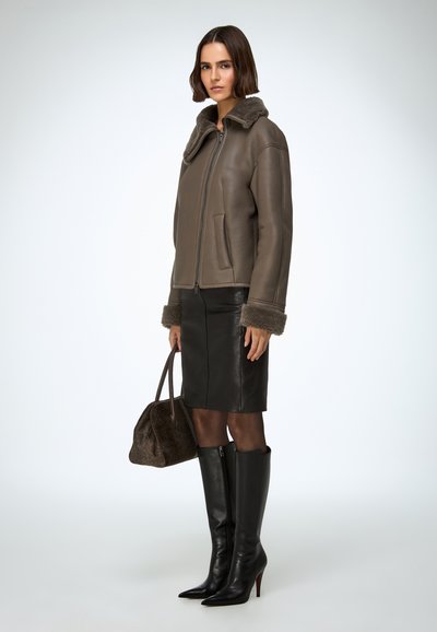 Hnedá kožená bunda s kožúškovým golierom, skombinovaná s čiernou koženou sukňou, čiernymi vysokými čižmami na podpätku po kolená a hnedou kabelkou.