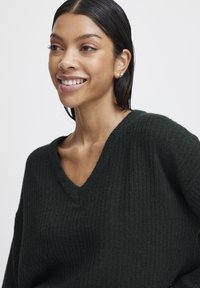 Pull en maille côtelée vert foncé avec un col en V, fabriqué dans un matériau doux. Il a des manches longues et une coupe décontractée.