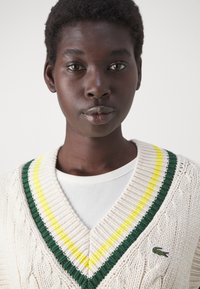 Cardigan en tricot crème avec décolleté en V, présentant des bords rayés verts et jaunes. Texture en relief à torsades et logo brodé sur la poitrine.