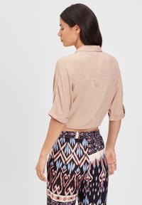 Blusa beige a maniche corte con risvolti e dettaglio dei bottoni sul retro, abbinata a pantaloni neri e colorati con motivi geometrici.