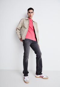 Chaqueta ligera beige, camiseta rosa, vaqueros oscuros grises y zapatillas blancas con rayas rojas. El modelo posa con una mano en la cadera frente a un fondo sencillo.