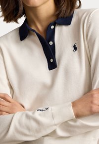 Polo blanc à manches longues avec col marine, doté d'une patte de boutonnage à trois boutons, en tissu texturé, et avec un logo brodé sur la poitrine et la manche.