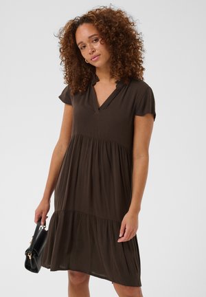 Femme aux cheveux bouclés portant une robe marron ample à manches courtes, tenant un sac à main noir sur un fond uni.