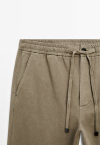 Khaki joggingové kalhoty s elastickým pasem, stahovací šňůrkou a bočními kapsami. Hladká textura s uvolněným střihem a minimálním detailem.