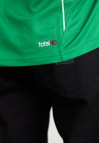 Zaļš acs sporta krekls ar baltām detaļām. Ar sarkano 'total90' logo. Kombinēts ar melnām biksēm, kurās redzamas kabatu šuvju detaļas.