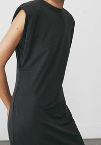 Musta minimalistinen mekko, jossa on lyhyet hihat, valmistettu sileästä kankaasta, rento malli ja huomaamattomat sivutaskut. Pehmeä materiaali ja tyylikäs muotoilu.