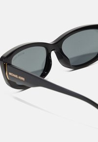 Juodos plastikinės saulės akiniai su pilkais lęšiais, turintys įlinkusią rėmelį ir elegantiškas šakas. Ant šono yra išgraviruotas "MICHAEL KORS".