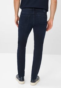 Jeans in denim blu scuro con una vestibilità slim, cinque tasche e dettagli di cucitura discreti sul retro, abbinati a scarpe in pelle nere.