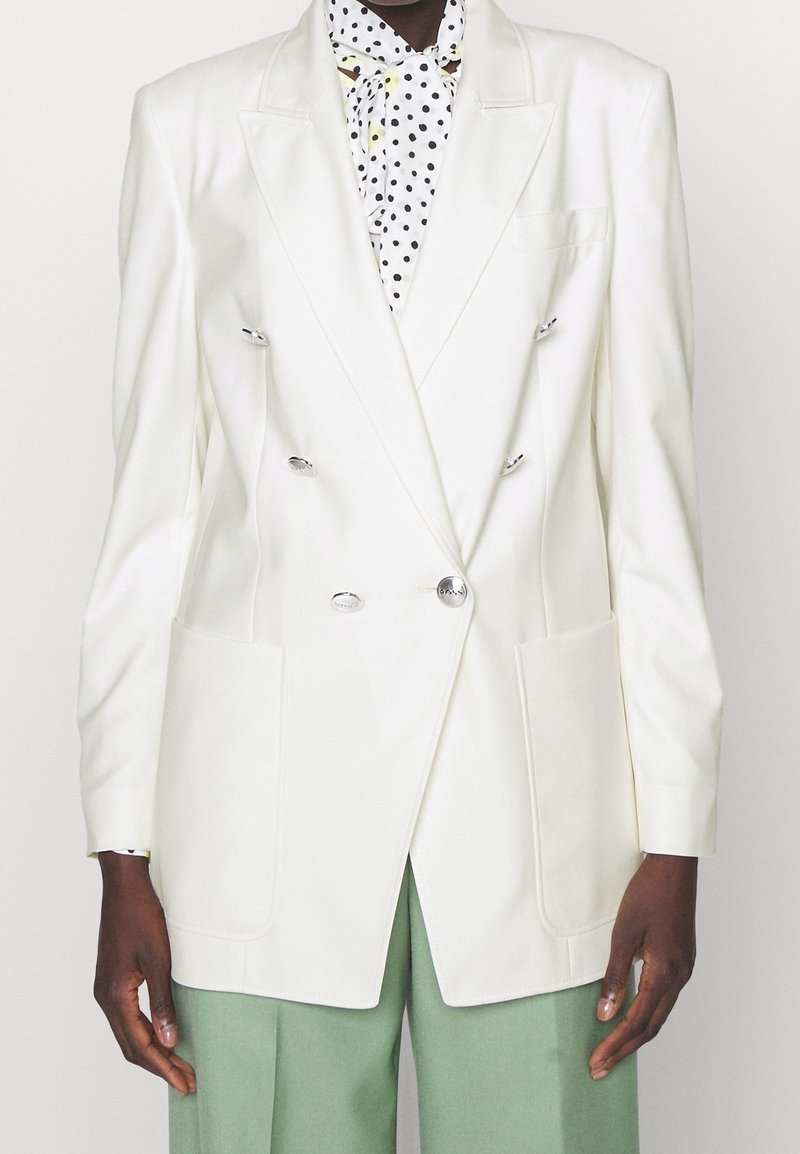 Blazer blanc à double boutonnage avec boutons argentés sur une blouse blanche à pois noirs, associé à un pantalon large vert clair.