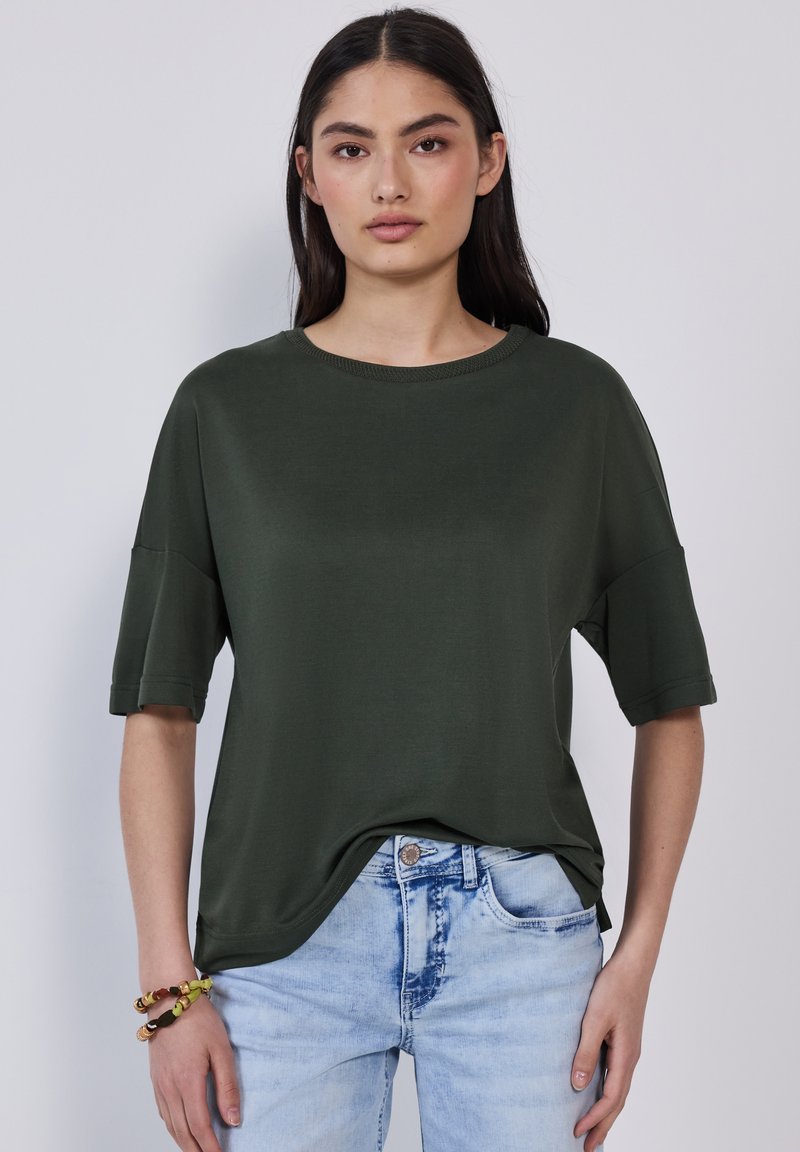 Tmavě zelené volné tričko s krátkým rukávem a kulatým výstřihem. Hladká textura, v kombinaci s světle modrými roztrhanými džíny.