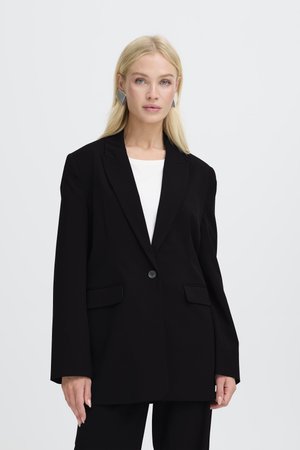 Femme blonde portant un blazer noir oversize sur un top blanc avec des boucles d'oreilles argentées géométriques, debout devant un fond clair uni.