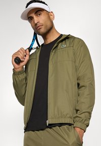 Veste de sport vert olive avec zip sur le devant, poignets et ourlet élastiques. T-shirt noir en dessous. Visière blanche et raquette de tennis à la main.