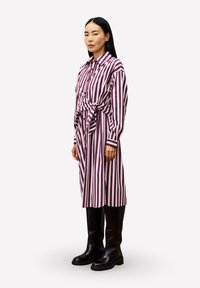 Frau in einem langärmeligen, lila und weißen, senkrecht gestreiften Kleid mit einer vorderen Schleife, kombiniert mit schwarzen kniehohen Stiefeln, steht seitlich.