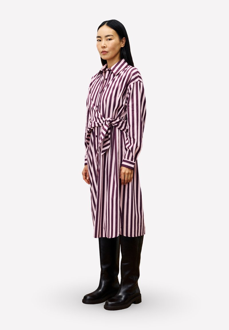 Frau in einem langärmeligen, lila und weißen, senkrecht gestreiften Kleid mit einer vorderen Schleife, kombiniert mit schwarzen kniehohen Stiefeln, steht seitlich.