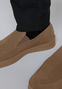 Barna, slip-on bőrcipő, puha velúrbőr textúrával, rugalmas oldalsó paneleknek köszönhetően és könnyű gumitalppal, fekete, szűkített nadrággal párosítva.