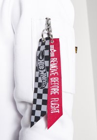 Deux porte-clés en tissu fixés à la fermeture éclair d'une poche de manche blanche ; l'un à carreaux noirs et gris avec l'inscription "Hot Wheels", l'autre rouge avec l'inscription "REMOVE BEFORE FLIGHT."