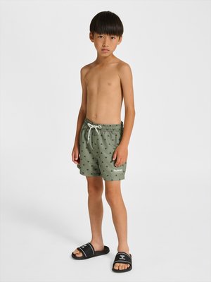 Dreng stående iført grønne mønstrede badeshorts og sorte slidesandaler mod en ensfarvet hvid baggrund.