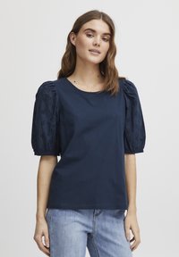 Chemise en coton bleu marine à manches courtes bouffantes avec des détails en dentelle. Encolure ronde, coupe décontractée, associée à un jean en denim bleu clair.