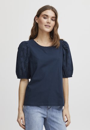 Marinblå bomullsskjorta med kort puffärm och spetsdetaljer. Rund halsringning, avslappnad passform, matchad med ljusblå denimjeans.