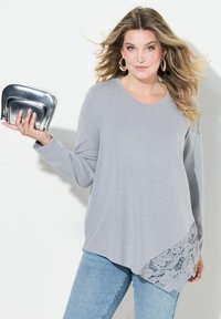 Grå langærmet top med V-udskæring, der har en asymmetrisk kant og blonde detaljer på den ene side. Parret med en metallisk sølv clutch.