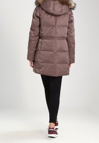 Brun puffjacka med huva kantad med päls, åtstramad midja och horisontell quiltning. Bärs över svarta leggings och matchas med mörka sportskor.