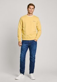 Felpa gialla con scollo rotondo, con colletto bianco e logo sul petto, abbinata a jeans blu e scarpe da ginnastica bianche. Modello in posizione frontale.