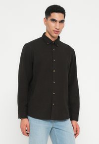 Hombre vistiendo una camisa negra abotonada con collar y jeans de color azul claro, de pie frente a un fondo blanco liso.